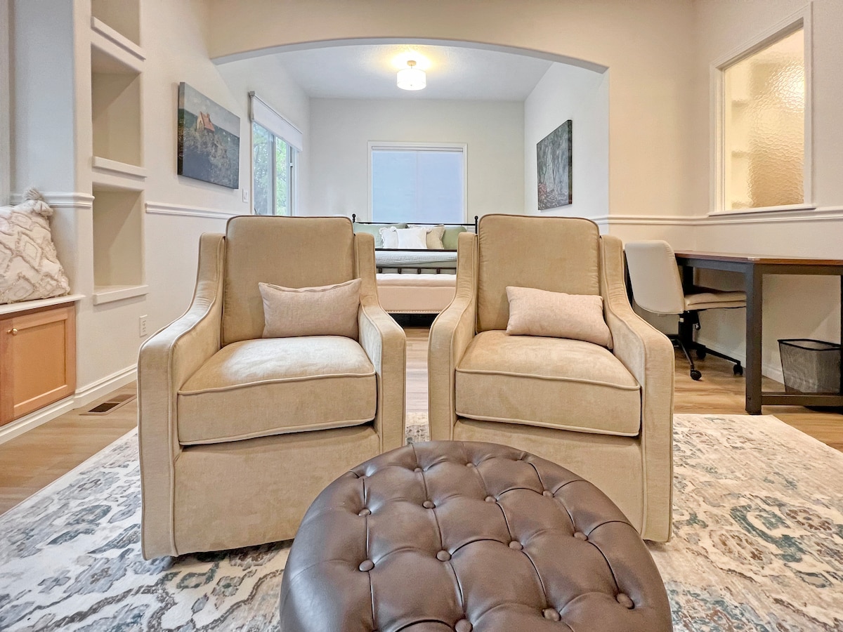 Two beige upholstered chairs are positioned facing each other on a patterned rug, creating a comfortable seating area. A round, tufted leather ottoman sits in the center. Natural light filters through large windows, illuminating the space that leads into an adjoining room.
