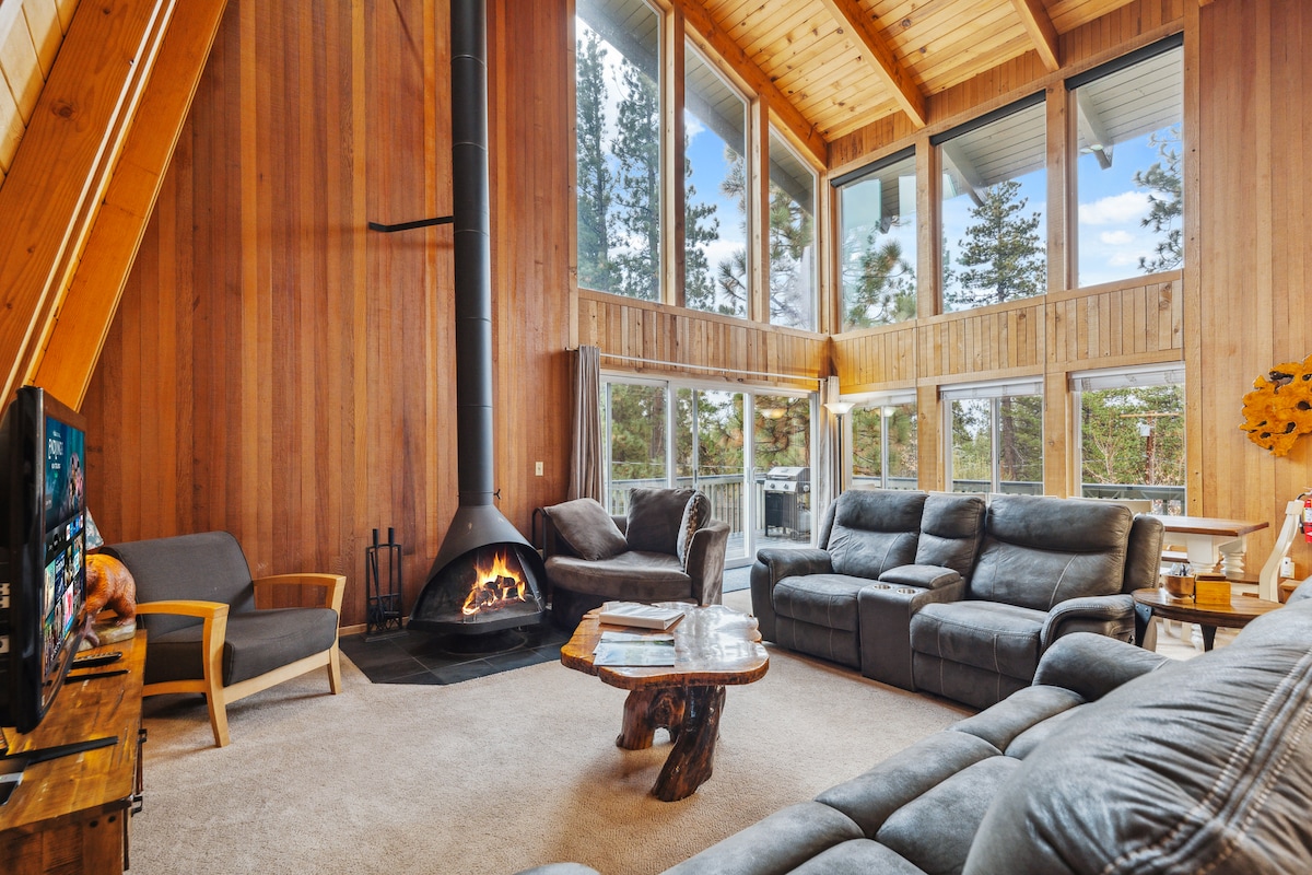 A spacious living area features high vaulted ceilings and large floor-to-ceiling windows that provide ample natural light and views of the surrounding forest. Comfortable seating includes both a reclining sofa and armchairs, paired with a rustic wooden coffee table and a stylish fireplace.