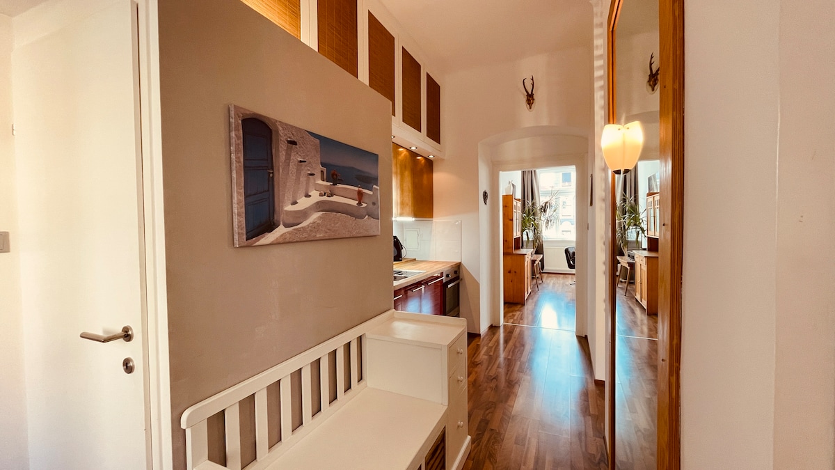 A welcoming hallway is presented, featuring a white bench and a decorative photograph on the wall. The space leads toward a bright living area, with wooden flooring and ample natural light coming from the open doorways.
