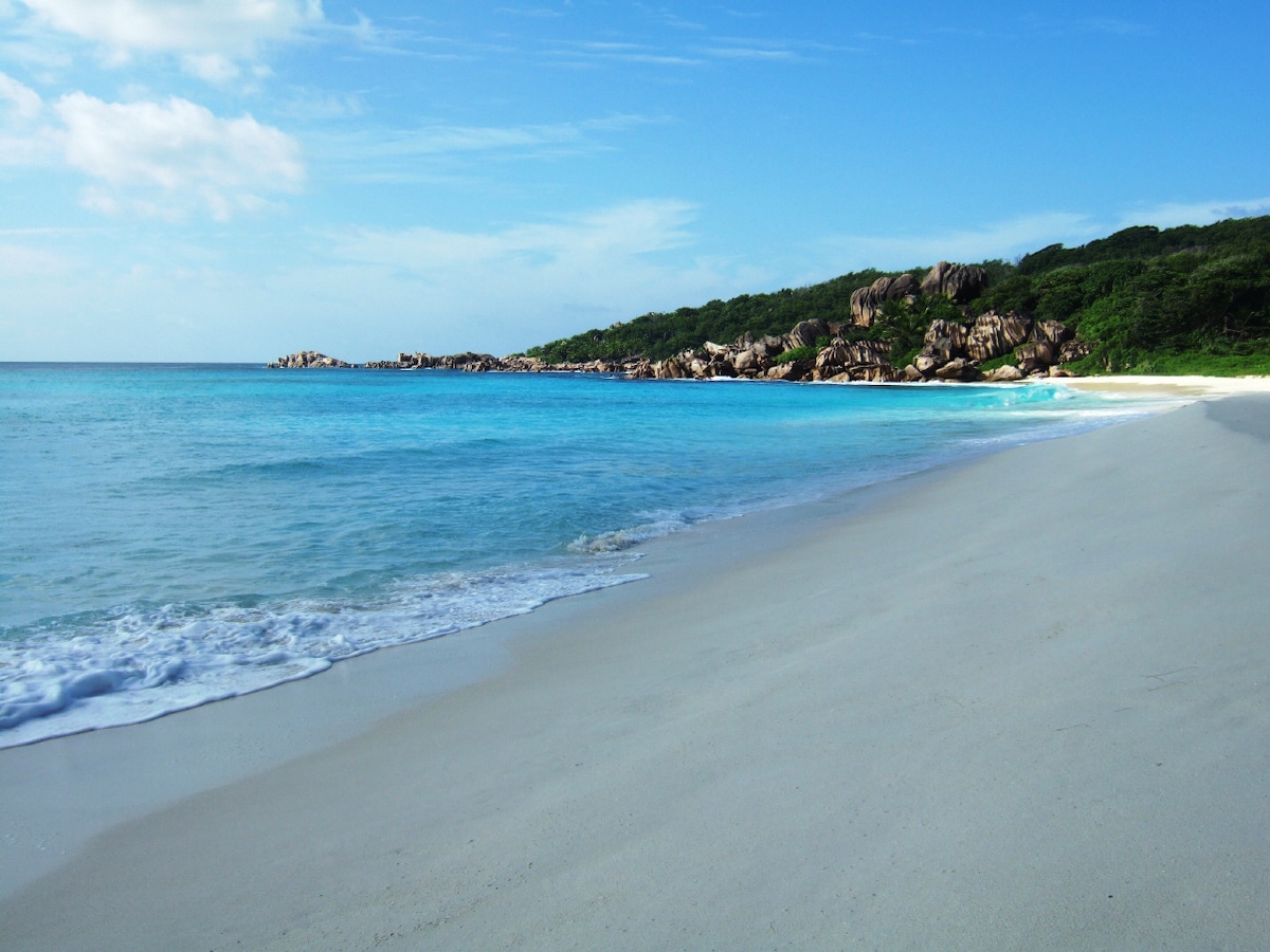 A serene beach scene is captured, featuring soft white sand meeting clear turquoise waters. Gentle waves lap at the shoreline, with lush greenery and rocky formations visible in the background under a bright blue sky. The tranquil setting invites relaxation and exploration.
