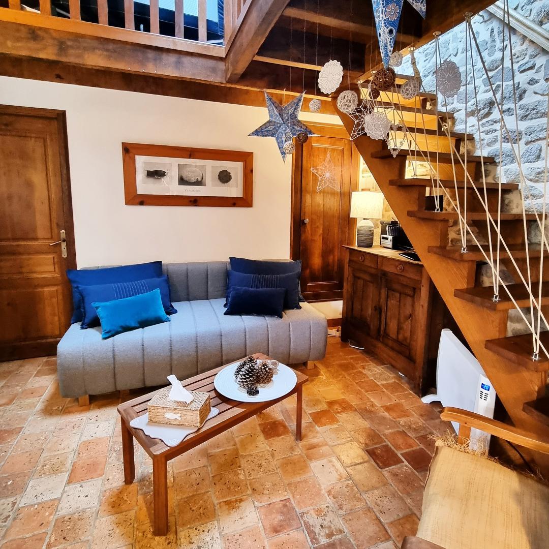 A welcoming living area is displayed, featuring a light grey sofa adorned with blue accent pillows. A wooden coffee table is centered on the terracotta tile floor, complemented by decorative elements. The space is brightened by warm lighting, with wooden elements and a staircase visible in the background.