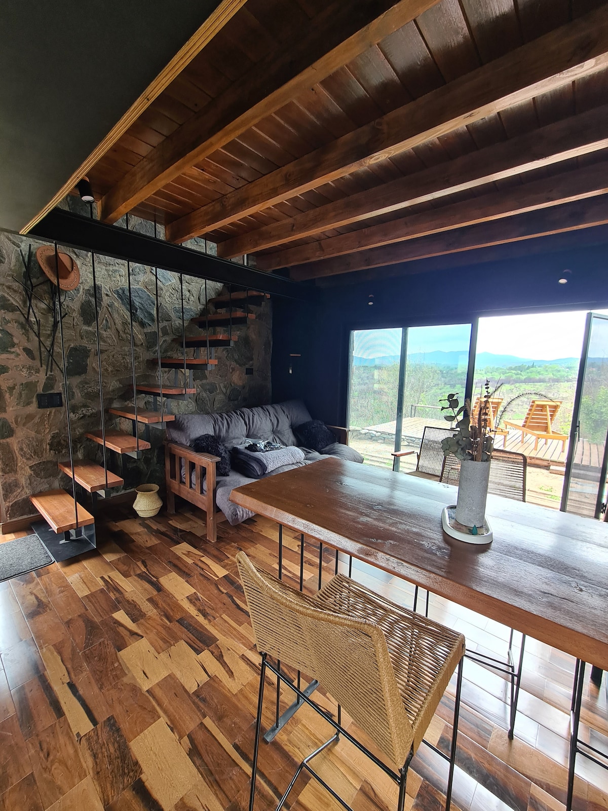 A spacious living area is illuminated by natural light, featuring a wooden ceiling and a stone accent wall. A comfortable sofa is positioned near large glass doors that open to expansive views, with a dining table and rustic chairs enhancing the cozy ambiance.