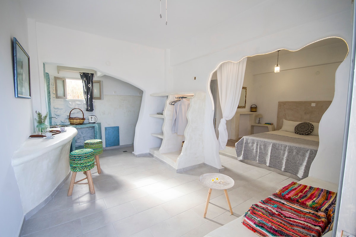 An open-plan living space features a cozy sleeping area with a spacious bed and decorative throw. A kitchenette is equipped for meal preparation, while a seating arrangement of colorful stools invites relaxation. Natural light enters through strategically placed windows, complementing the minimalist design.