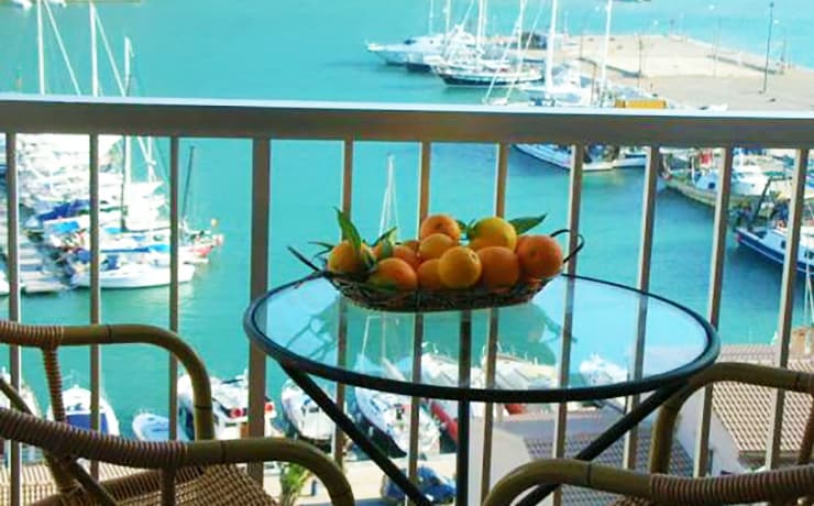 A balcony view overlooking the Mediterranean Sea features a glass table topped with a basket of oranges. In the background, boats are moored in the harbor, with calm turquoise waters reflecting the vibrant colors of the scene.