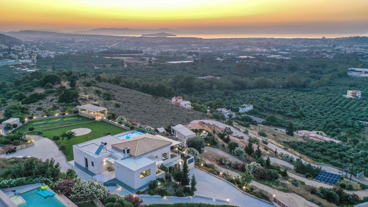 An elevated view captures Villa Kedria and its surroundings at sunset. The villa is situated on a hillside, surrounded by lush greenery and a private pool, with expansive views of the ocean and the charming town of Chania in the distance.