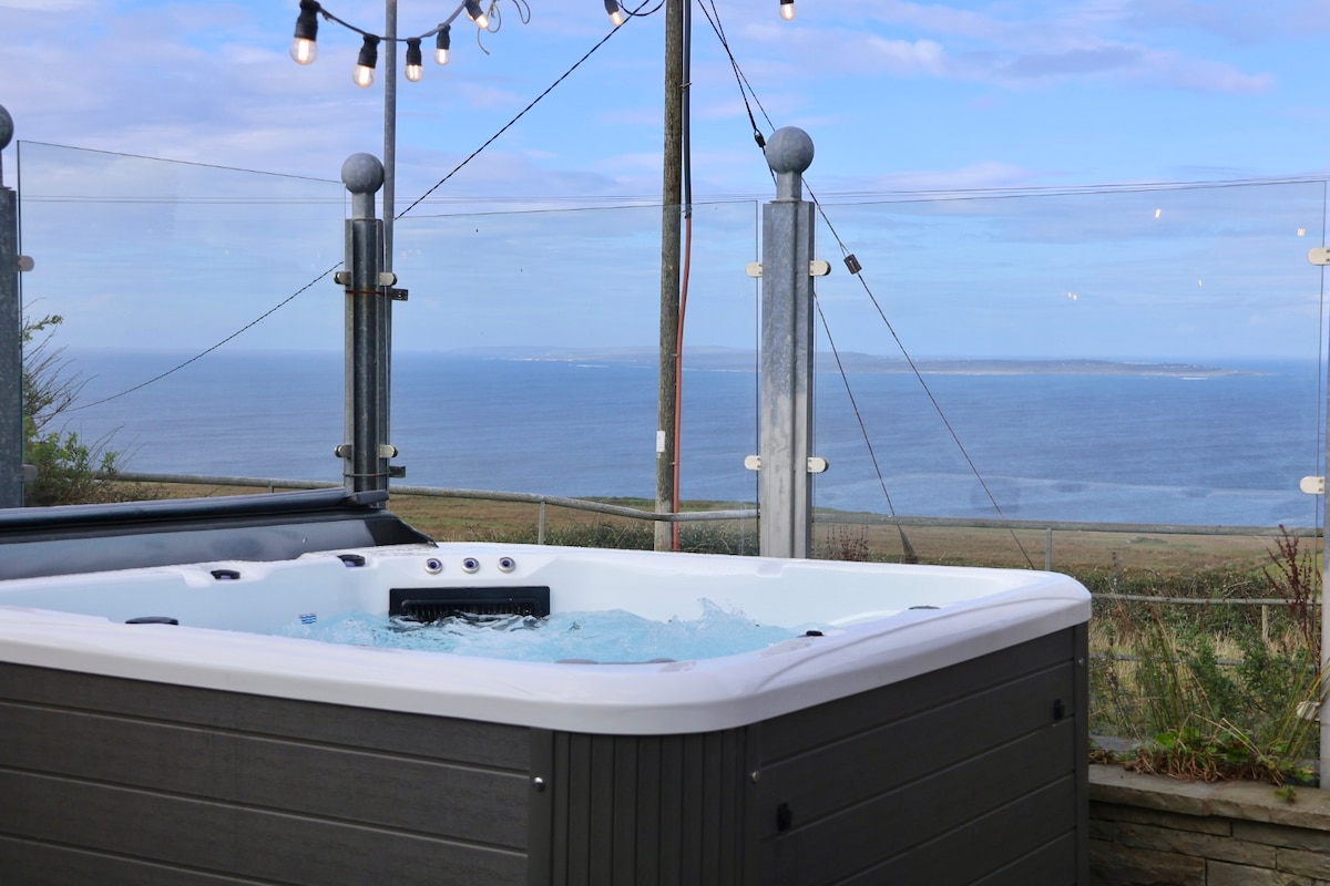 A hot tub awaits relaxation with bubbles creating gentle waves on the surface. Glass panels provide an unobstructed view of the sea, while string lights add subtle illumination above. The expansive horizon reveals tranquil waters and distant islands.