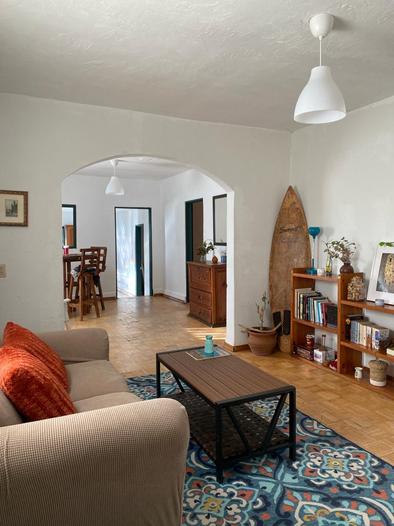 A comfortable living area features a beige sofa adorned with orange pillows, positioned beside a low coffee table. Colorful rugs add warmth to the space, which is complemented by wooden furniture and decorative plants. An archway leads to the dining area in the background.
