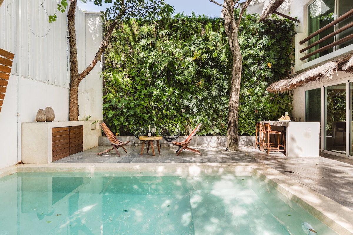 A serene outdoor area features a private, unheated pool surrounded by tropical greenery. Two wooden lounge chairs are positioned beside a wooden table, offering a comfortable space for relaxation. Sunlight filters through the leaves, creating dappled patterns on the stone patio.