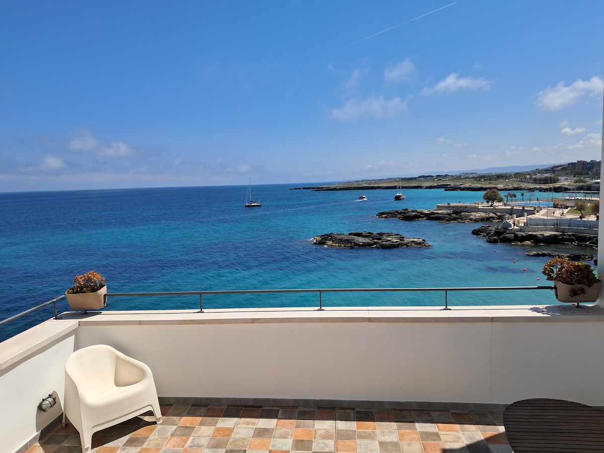 A balcony offers a stunning view of the clear blue sea, showcasing gentle waves and distant sailboats. Potted plants add a touch of greenery to the space. The expansive horizon blends seamlessly where the water meets the sky under soft, fluffy clouds.