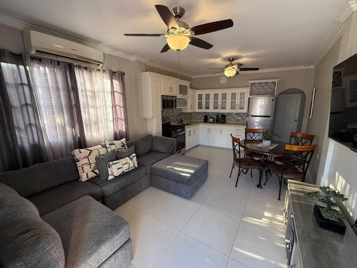 A spacious living and dining area features a large gray sectional sofa and a dining table with four chairs. Natural light filters through sheer curtains, illuminating the modern kitchen with white cabinetry and stainless steel appliances. A ceiling fan provides additional comfort.