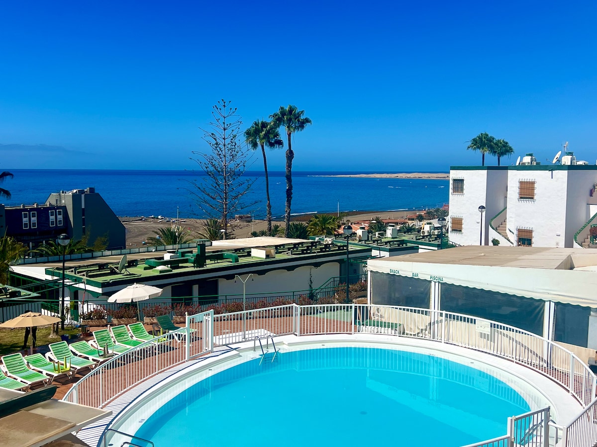 A circular swimming pool is centrally located, surrounded by sun loungers and umbrellas. Lush palm trees frame the scene, with the clear blue ocean visible in the background under a bright sky. Nearby buildings are partially visible, complementing the tranquil space.