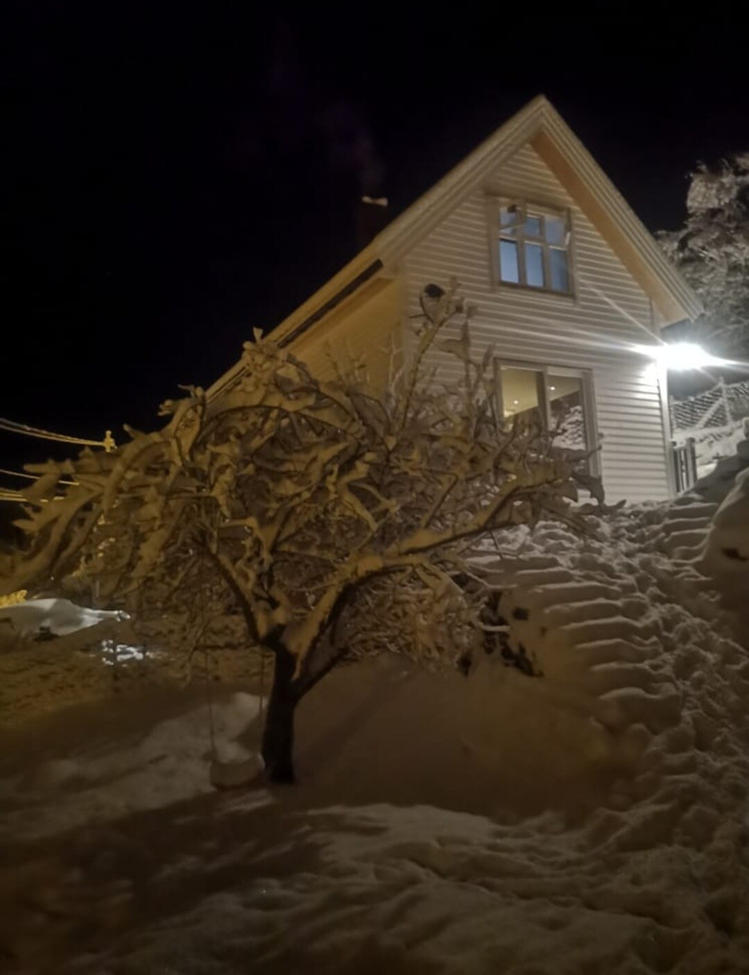 A charming white house is illuminated by a warm outdoor light, showcasing a snowy landscape. A snow-covered tree is positioned near the steps leading up to the entrance, creating a serene winter scene during nighttime.
