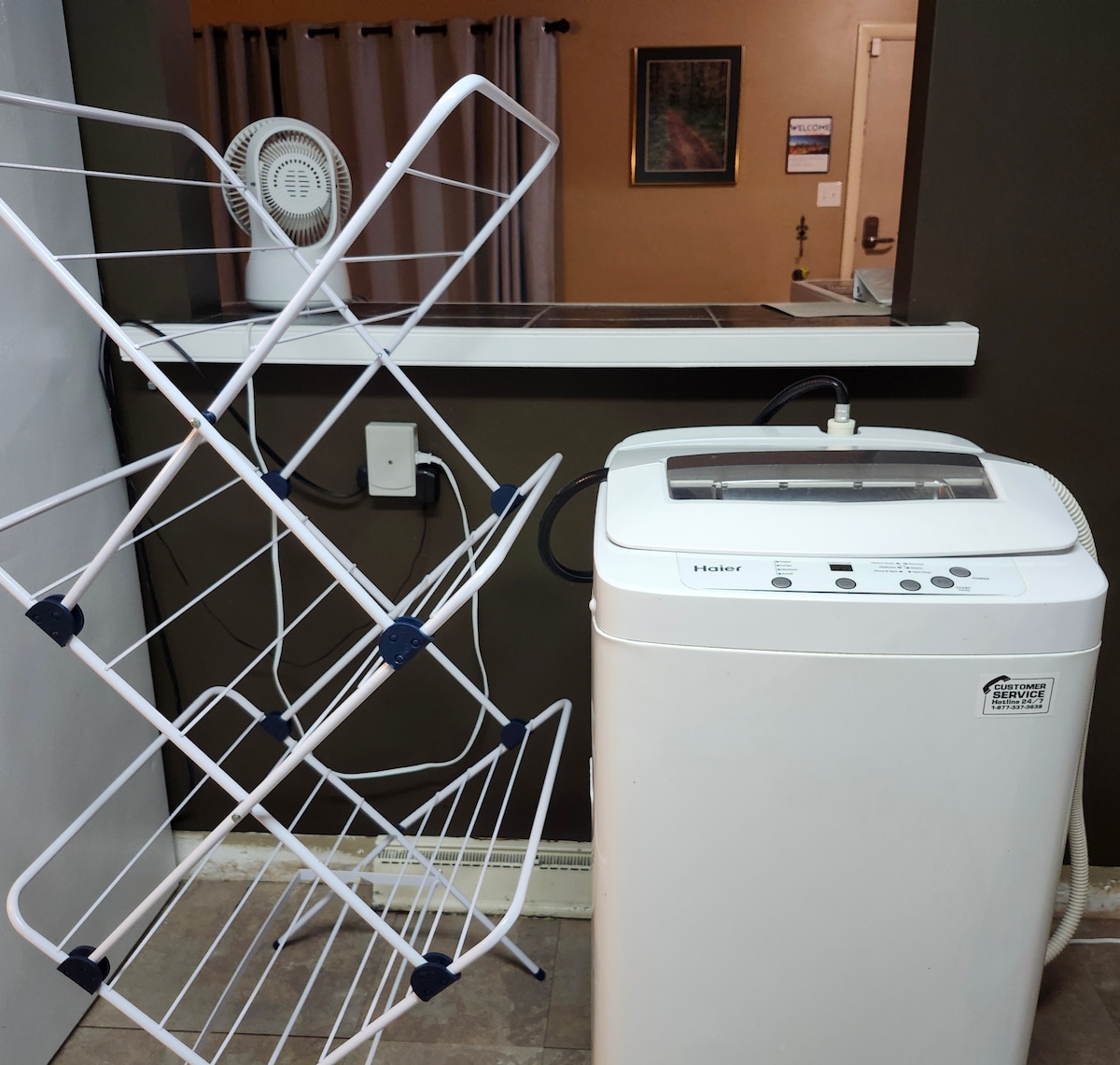A portable washing machine is positioned next to a drying rack, which is set to hold laundry. The machine features a control panel on top with various cycle options. The wall behind is painted in a muted brown tone, with a glimpse of a doorway partially visible.