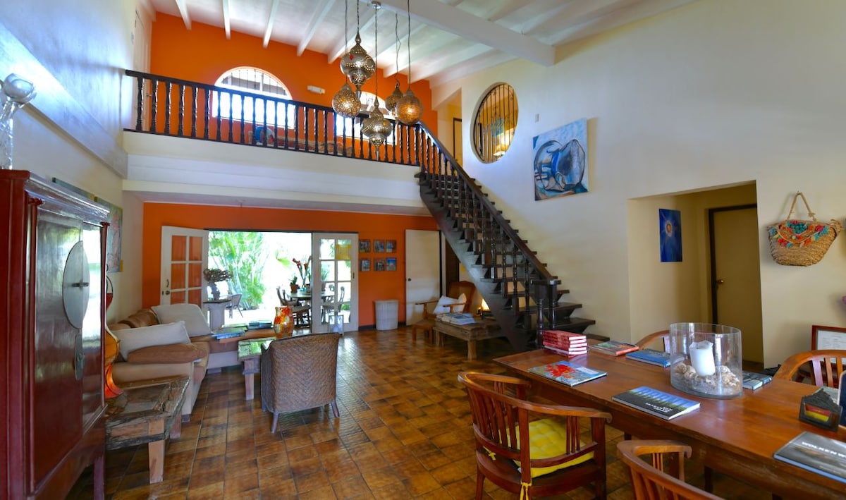 A spacious communal area is highlighted by high ceilings and wooden beams. Comfortable seating arrangements feature a mix of sofas and chairs, while a large dining table occupies the center. A staircase leads to an upper level, and tropical greenery is visible through the large windows.
