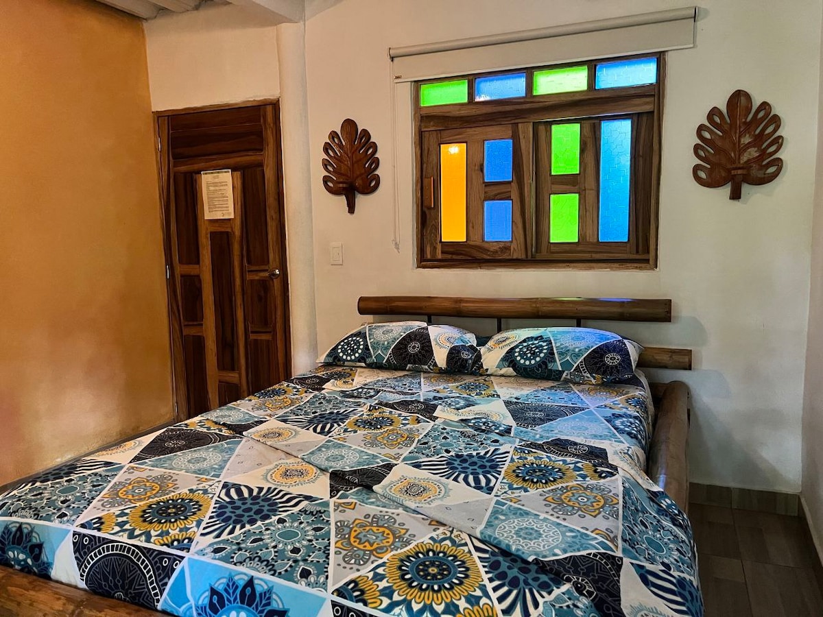 A spacious bedroom features a large bed adorned with a patterned quilt. A colorful window with panes of blue, green, and orange allows natural light to brighten the room. Decorative wooden elements complement the warm tones of the walls and furnishings.