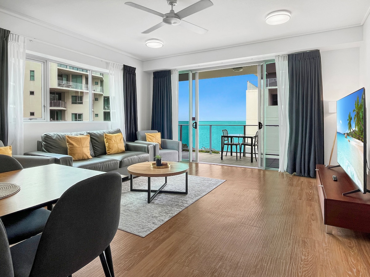 The spacious living area features soft seating with accent pillows, hardwood flooring, and a large smart TV. Large windows and sliding doors frame scenic ocean views, while a dining table for four invites shared meals. Natural light fills the space, creating a bright atmosphere.