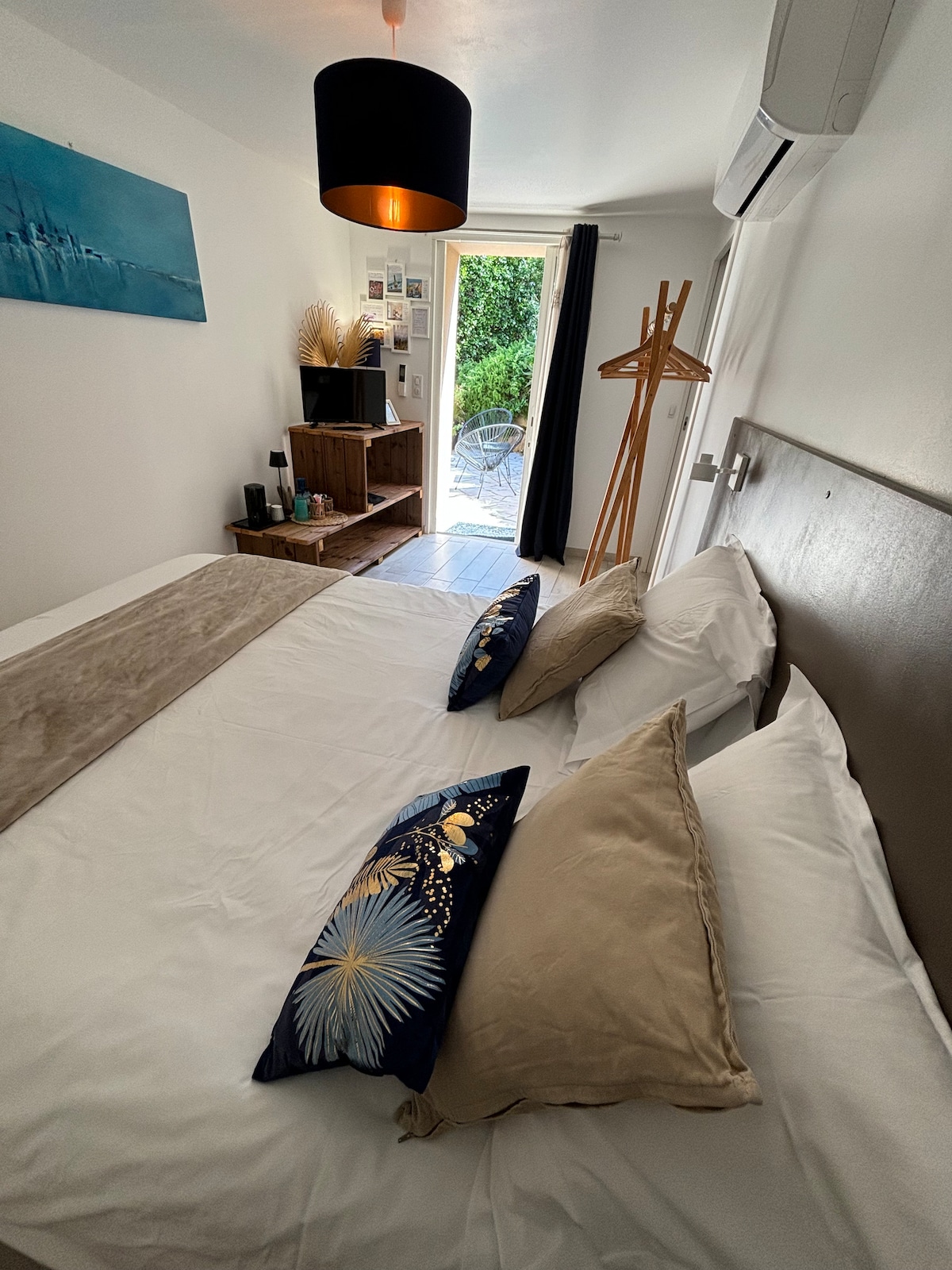 The room features a spacious bed adorned with decorative pillows and a light throw. A small wooden unit provides storage, while a decorative hat stand offers additional functionality. A bright door leads to an outdoor area, allowing for natural light to fill the space.
