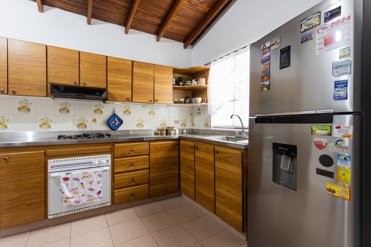A warm kitchen area features wooden cabinetry complemented by stainless steel appliances. A gas stove and sink are situated along the countertop, providing ample workspace. A refrigerator adorned with magnets stands nearby, while a window lets in natural light, enhancing the inviting environment.