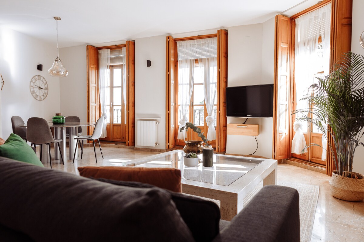 A bright living area features multiple windows with wooden frames, allowing natural light to illuminate the space. A comfortable sofa faces a wall-mounted flat-screen TV, while a dining table with modern chairs provides a cozy eating area. Decorative plants enhance the inviting atmosphere.