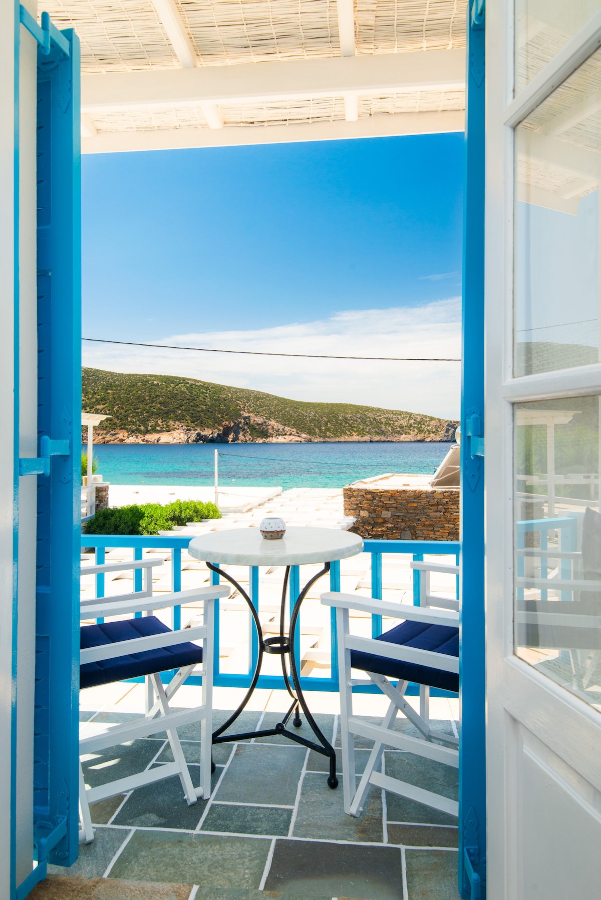Open doors reveal a small balcony with a table and two chairs, overlooking the serene coastline. Vibrant blue accents frame the view of the beach and distant hills under a clear sky, inviting relaxation and tranquility.