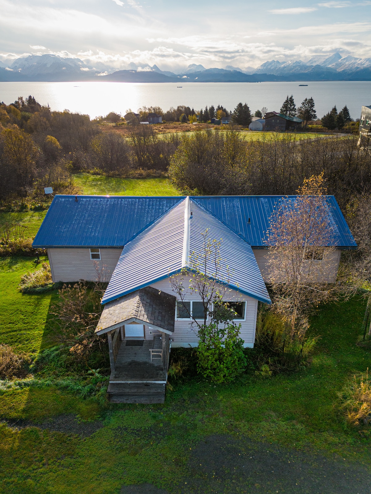 An aerial view reveals a spacious home with a blue metal roof and a welcoming entrance. Surrounding greenery and trees lead to scenic water views, with mountains in the distance. The well-kept exterior reflects a private feel amidst the natural landscape.