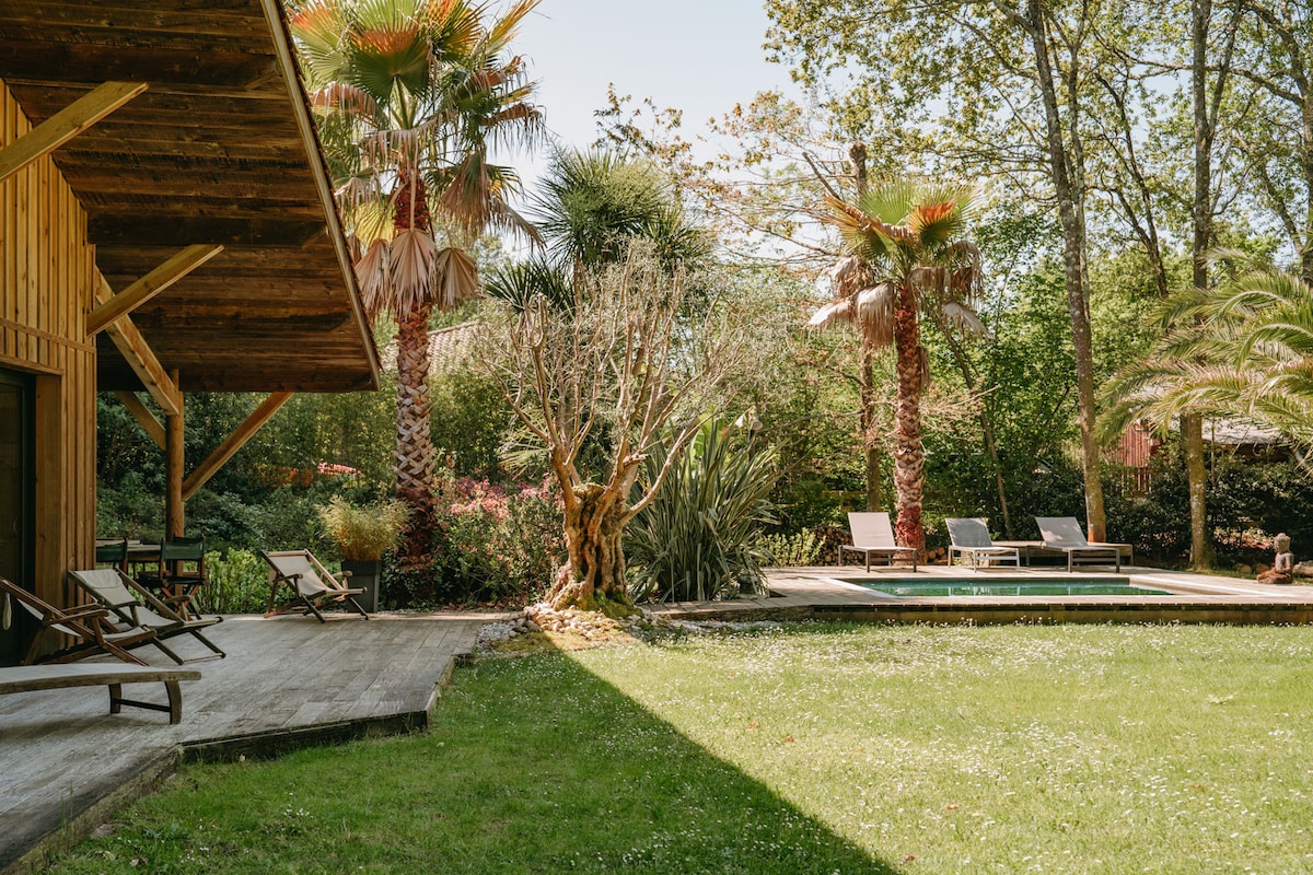 A wooden structure is showcased, featuring a covered porch with loungers. A lush green lawn extends towards an outdoor pool area, surrounded by tropical palm trees and neatly arranged lounge chairs. Natural light filters through the trees, enhancing the serene outdoor space.
