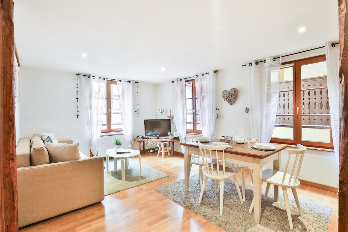 A spacious living area features large windows allowing natural light, with a comfortable sofa and an entertainment setup. A dining table with four chairs is positioned near the kitchen, complemented by decorative elements on the walls, creating an inviting yet functional space.