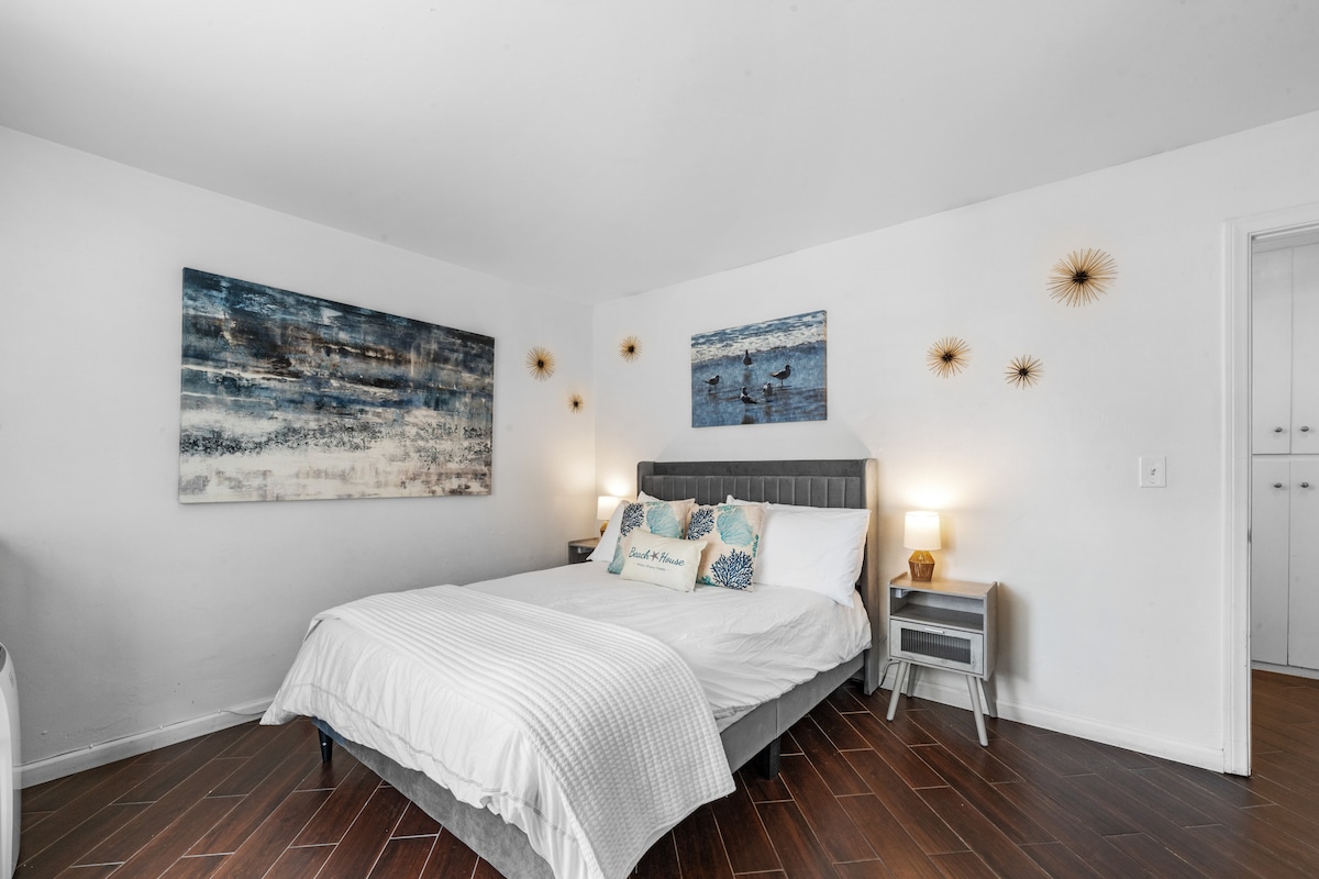 Svetlá priestranná spálňa so sivým rámom postele a bielou posteľnou bielizňou. Na posteli sú 4 vankúše a tri dekoratívne vankúše. Dve plážové lampy sú zapnuté a miestnosť je jasne osvetlená. 
