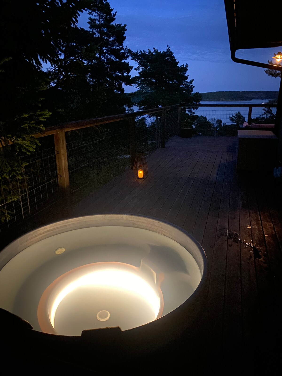 A hot tub is situated on a wooden deck, softly illuminated by integrated lighting. The serene view of the water is visible in the background, framed by surrounding trees. A lantern adds a touch of ambiance to the peaceful evening setting.