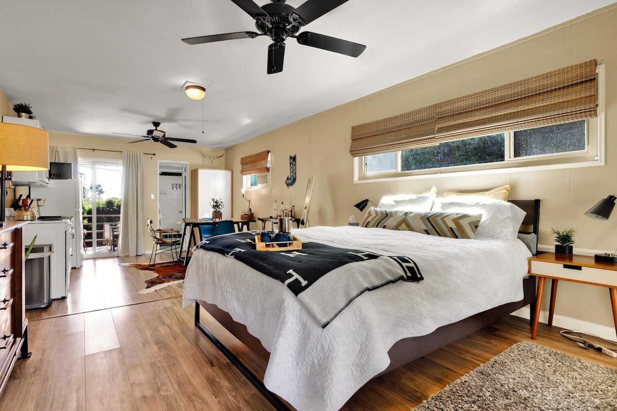 A spacious studio unit features a large bed adorned with crisp white linens. Natural light fills the room through two windows. A kitchenette is visible to the left, and a dining area is set against the wall. A cozy area rug adds warmth to the wooden floor.