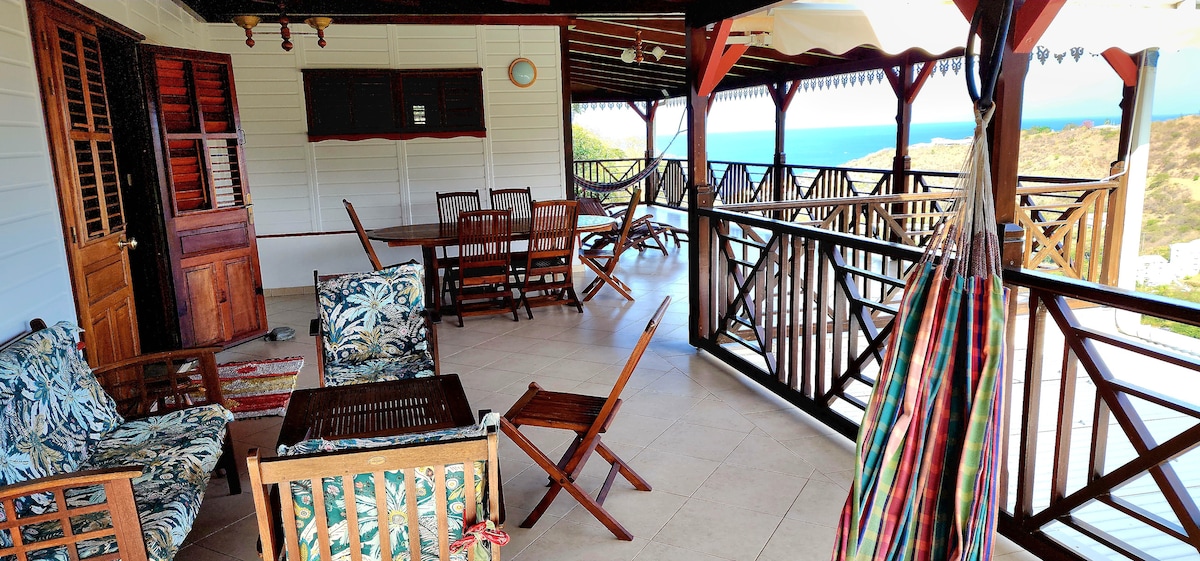 An outdoor terrace features wooden furniture, including a dining table with six chairs and two lounge chairs. A vibrant hammock is displayed along one side, and the sea can be seen in the distance through the balustrade. Ceiling fans are installed for comfort.