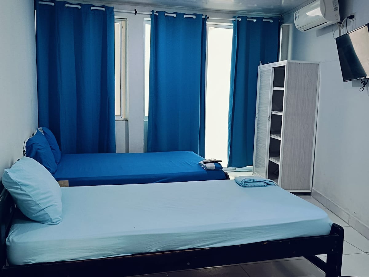 The room features two beds with light blue bedding, positioned near windows draped with dark blue curtains. A white cabinet stands against one wall, while a television is mounted nearby. Natural light enters through the partially open windows.