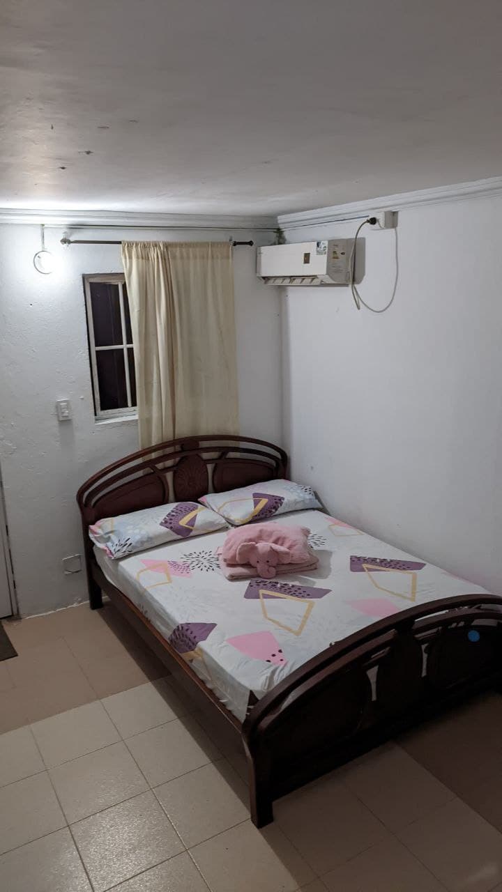 A cozy sleeping area features a wooden bed with a colorful patterned sheet set. A soft pillow is placed near the headboard, and a window with light curtains allows gentle light to enter. An air conditioning unit is mounted on the wall.