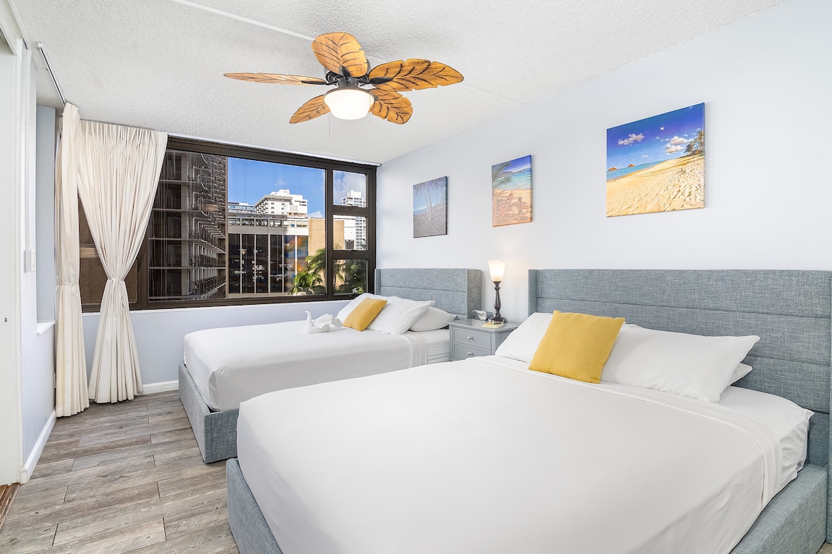 A bright bedroom features two spacious queen-size beds with light bedding and yellow accent pillows. Natural light enters through a large window, complemented by sheer curtains. Colorful beach-themed artwork adorns the walls, and a ceiling fan provides an inviting touch.