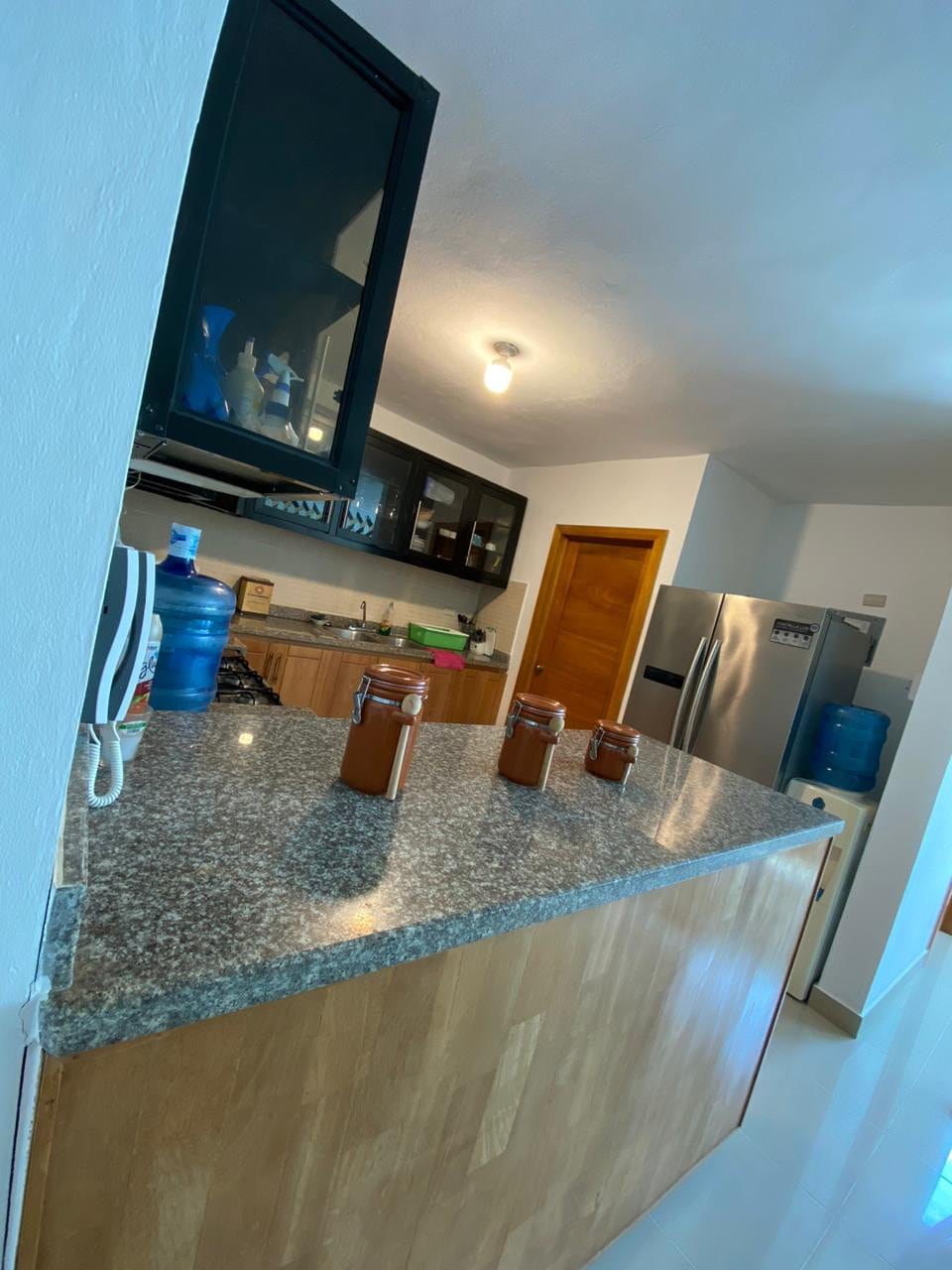 A modern kitchen features a large, polished countertop with several jars positioned along its edge. Upper cabinets, in dark tones, house dishes and cooking essentials, while a stainless-steel refrigerator and stove enhance the functional space. Natural light enters from a nearby door.