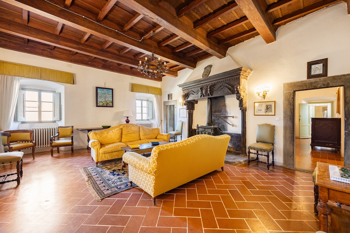 A spacious living room features a high wooden-beamed ceiling and terracotta tiled flooring. An antique monumental fireplace is situated against one wall, with a yellow sofa and various seating options arranged around it. Windows allow natural light to fill the room, enhancing its historical charm.