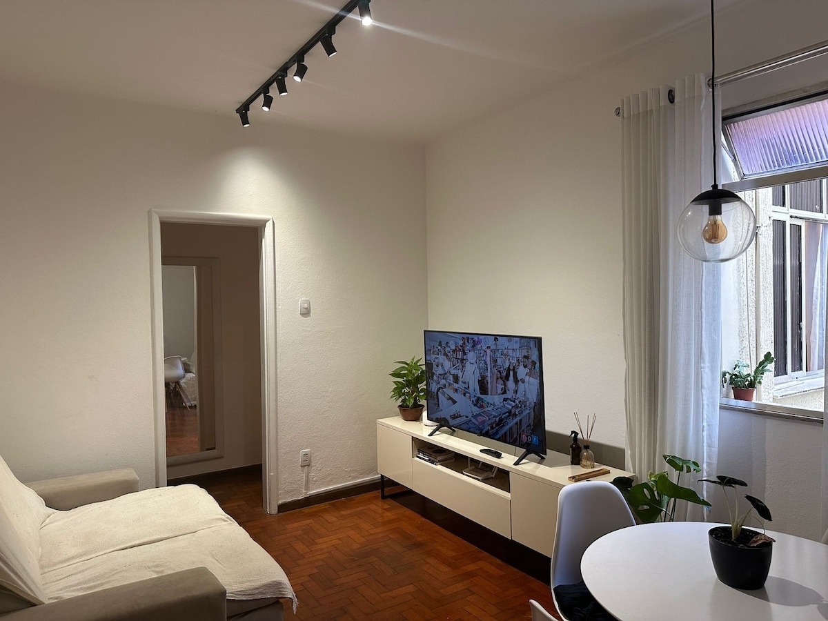 A well-lit living area features a white sofa and a mounted television on a light-hued media unit. Plants are positioned near the window, enhancing the space. A round dining table with minimalist chairs provides a welcoming spot for meals, complementing the room's cozy atmosphere.