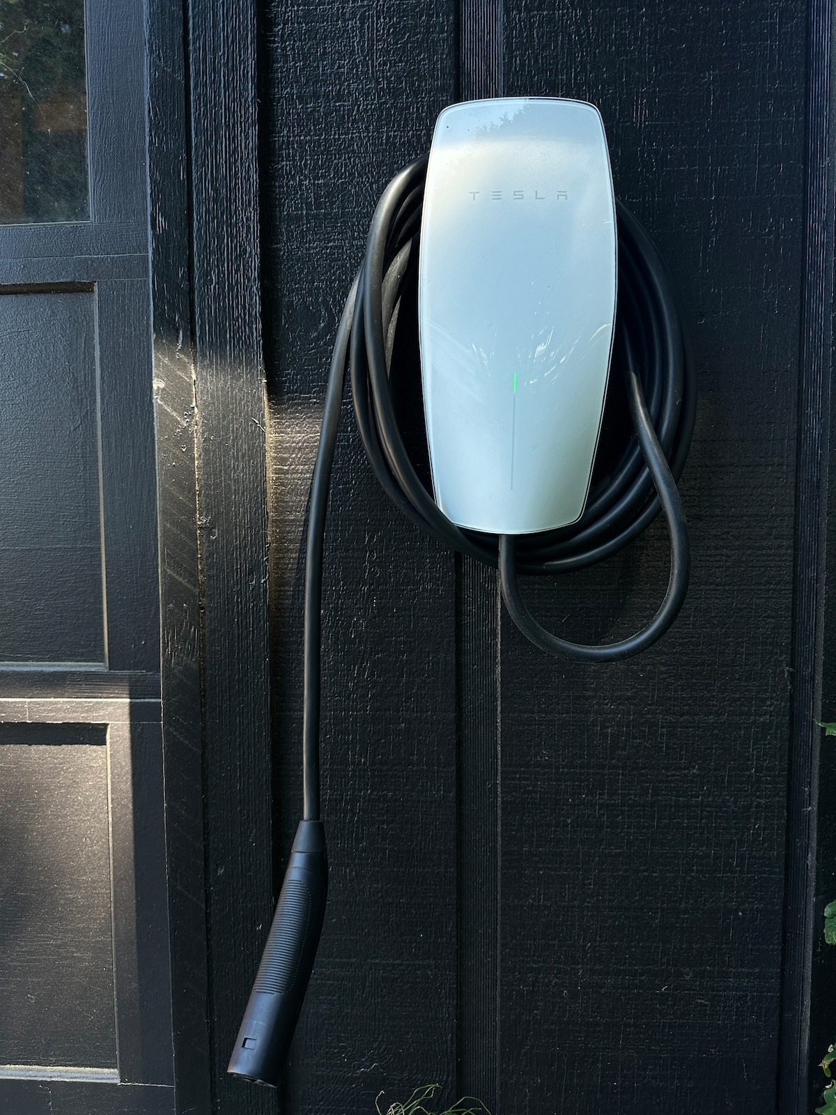 A Tesla charging station is mounted on a dark wooden wall. The sleek, white unit is connected to a long black charging cable, which hangs neatly beside it. Natural light accentuates the charging station, creating a clean and modern look.
