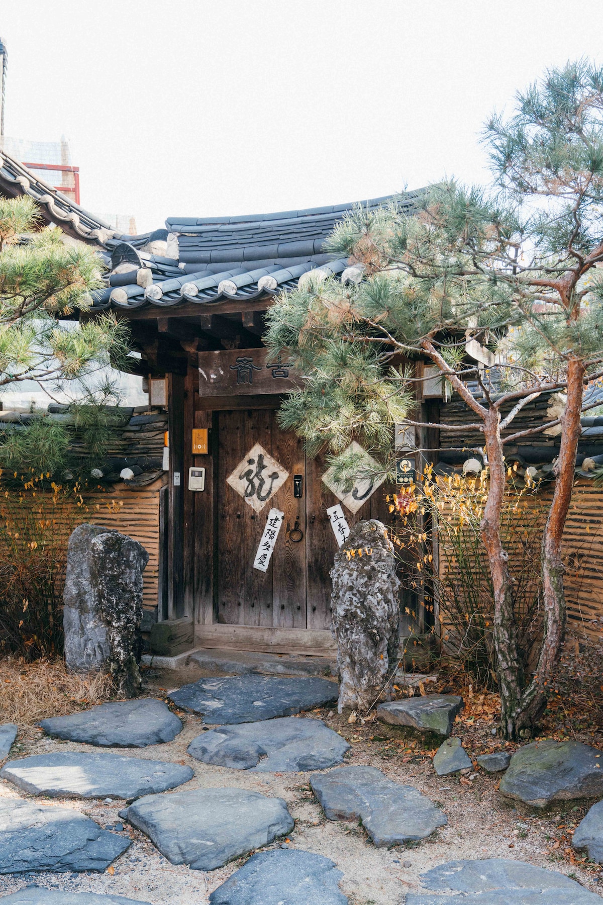 A traditional entrance gate with wooden doors is framed by natural stone pathways. Pine trees and decorative rocks enhance the serene atmosphere. The gate features traditional signage, complementing the historical architecture of the surrounding area.