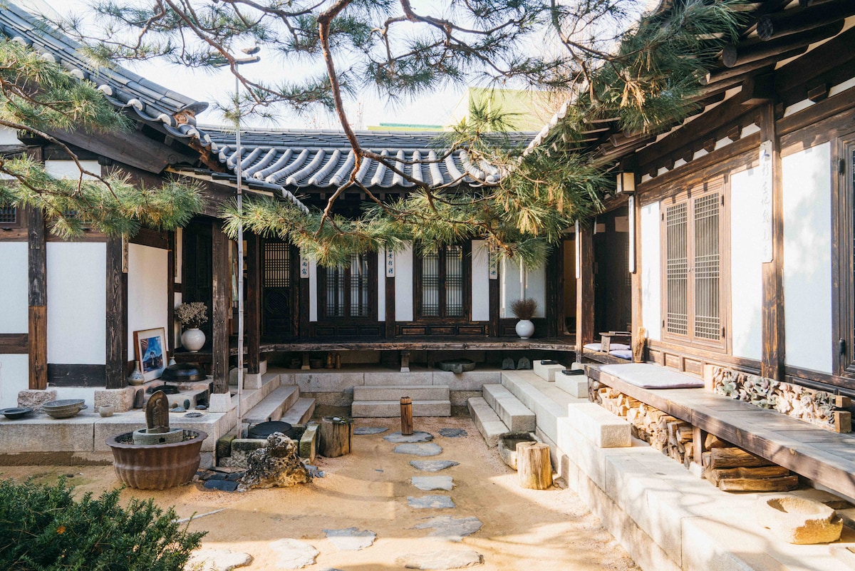A traditional Hanok courtyard is framed by wooden structures, featuring a stone path leading through a sandy area. Lush pine trees provide shade, while a wooden table sits alongside stone seating, inviting relaxation. Decorative elements such as an earthenware jar and small water basin enhance the space's cultural ambiance.