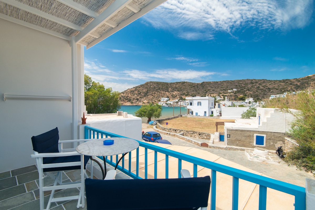 A balcony is shown overlooking a serene bay surrounded by hills. Outdoor furniture includes a small table and two chairs, with a scenic view of whitewashed buildings and greenery reflected in the calm water.