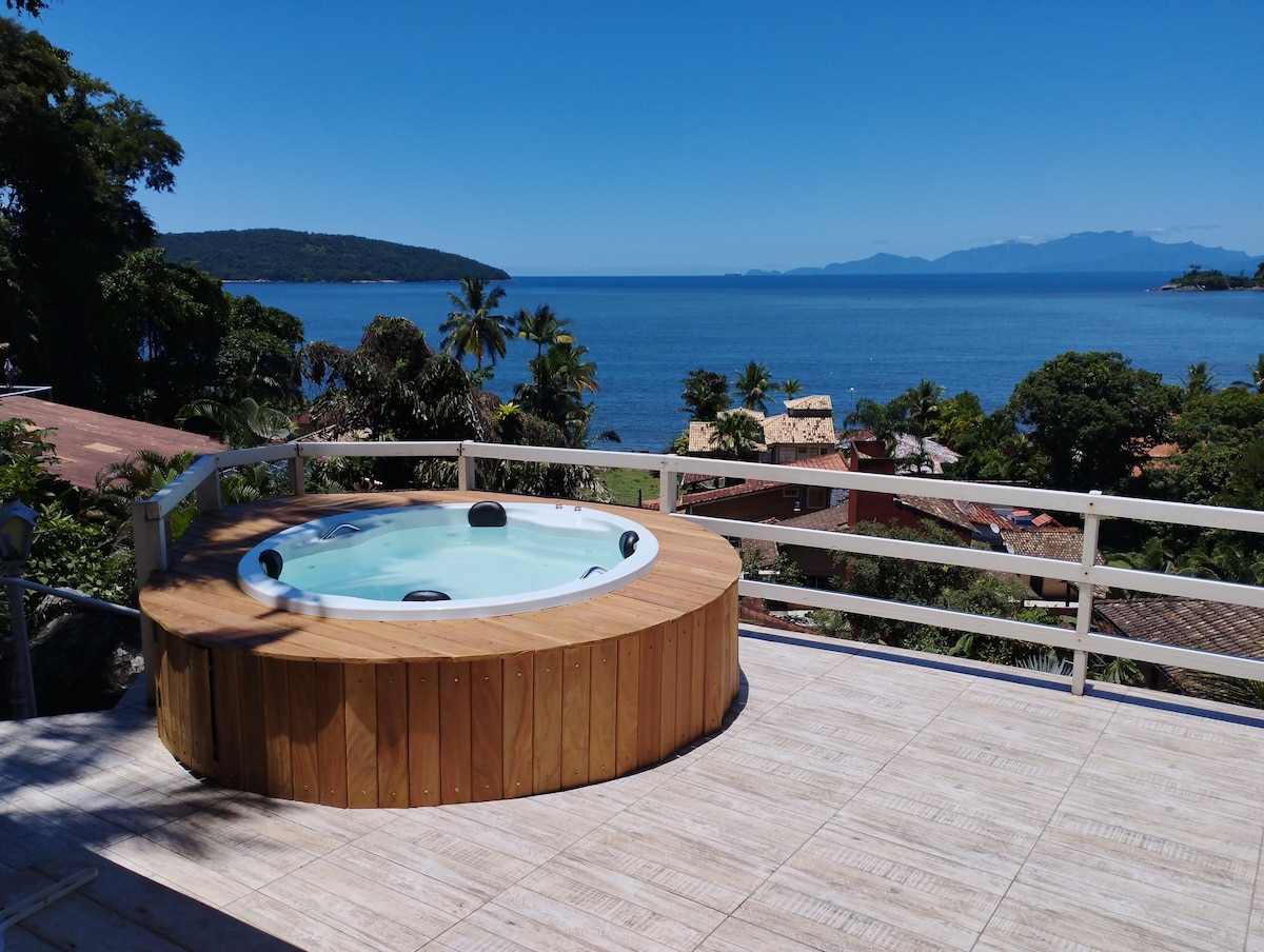 A hot tub encircled by wooden decking is positioned on a terrace, offering a clear view of the ocean. The scene is complemented by palm trees and rooftops in the foreground, with distant mountains visible on the horizon under a bright blue sky.