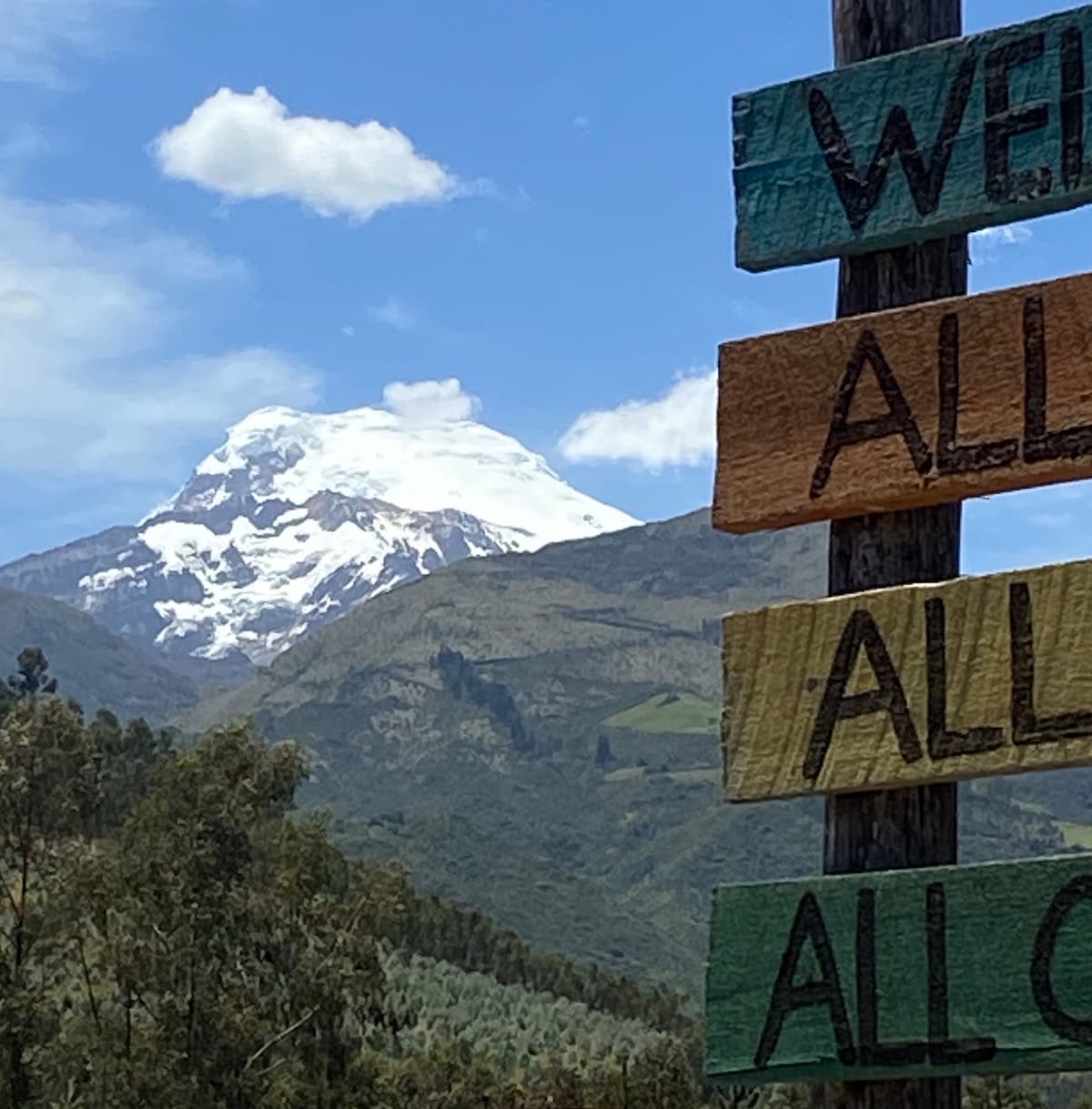 A vibrant wooden sign displaying the text 'WELCOME ALL' stands prominently in the foreground, with the majestic snow-capped Cayambe Volcano visible in the background. The lush green landscape stretches out beneath a clear blue sky, creating a tranquil and inviting scene.