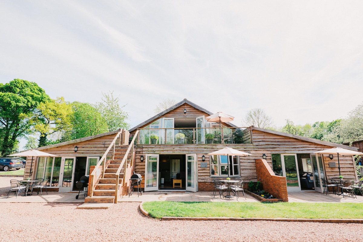 A spacious two-story cottage features a wooden façade, large glass windows, and multiple outdoor patio spaces. The entryway is flanked by parasols and outdoor seating, while a wooden staircase leads to the upper level. Lush greenery surrounds the property, contributing to a serene rural setting.