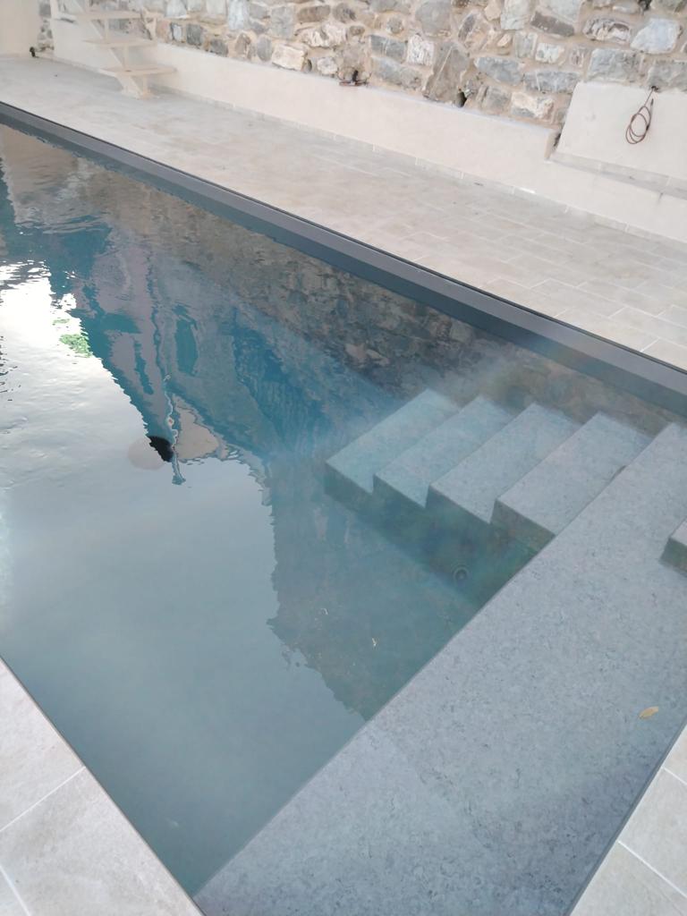 A serene swimming pool features a shallow entry with steps leading down into the clear water. The surrounding stone walls add a rustic touch, complementing the modern design of the pool area.