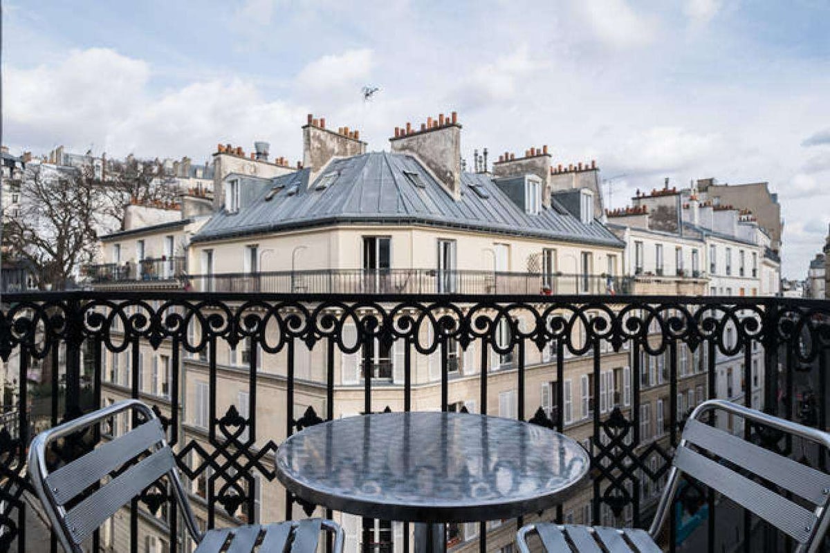 A small balcony is presented with a round metal table and two metal chairs. The ornate black railing frames a view of the neighboring buildings, showcasing their rooftop features and chimneys under a cloudy sky.
