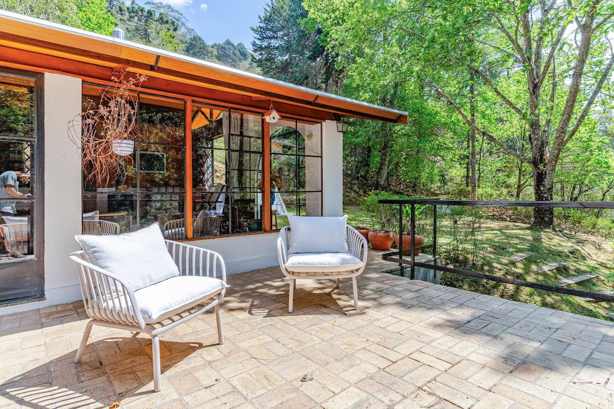 A serene outdoor terrace features two white lounge chairs positioned on a natural stone floor. Large windows and a sliding glass door lead into the interior, revealing a view of the surrounding greenery and sunlight filtering through the trees.