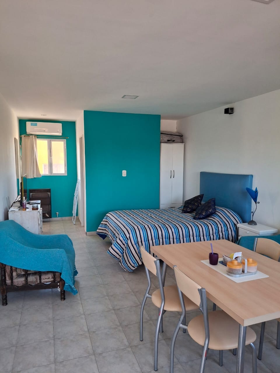 A modern apartment features a cozy sleeping area with a striped blanket draped over a spacious bed. A light blue accent wall complements a small dining table surrounded by four chairs, while a comfortable seating area is present in the corner beside a window.