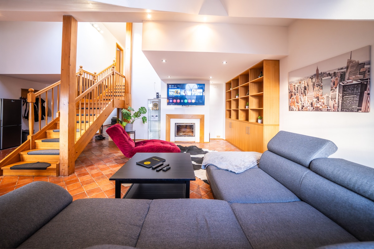 A modern living area is presented with a comfortable sectional sofa in grey facing a sleek coffee table. A large television is mounted on the wall, above a decorative fireplace. Natural light streams in, illuminating wooden features and the staircase in the background.