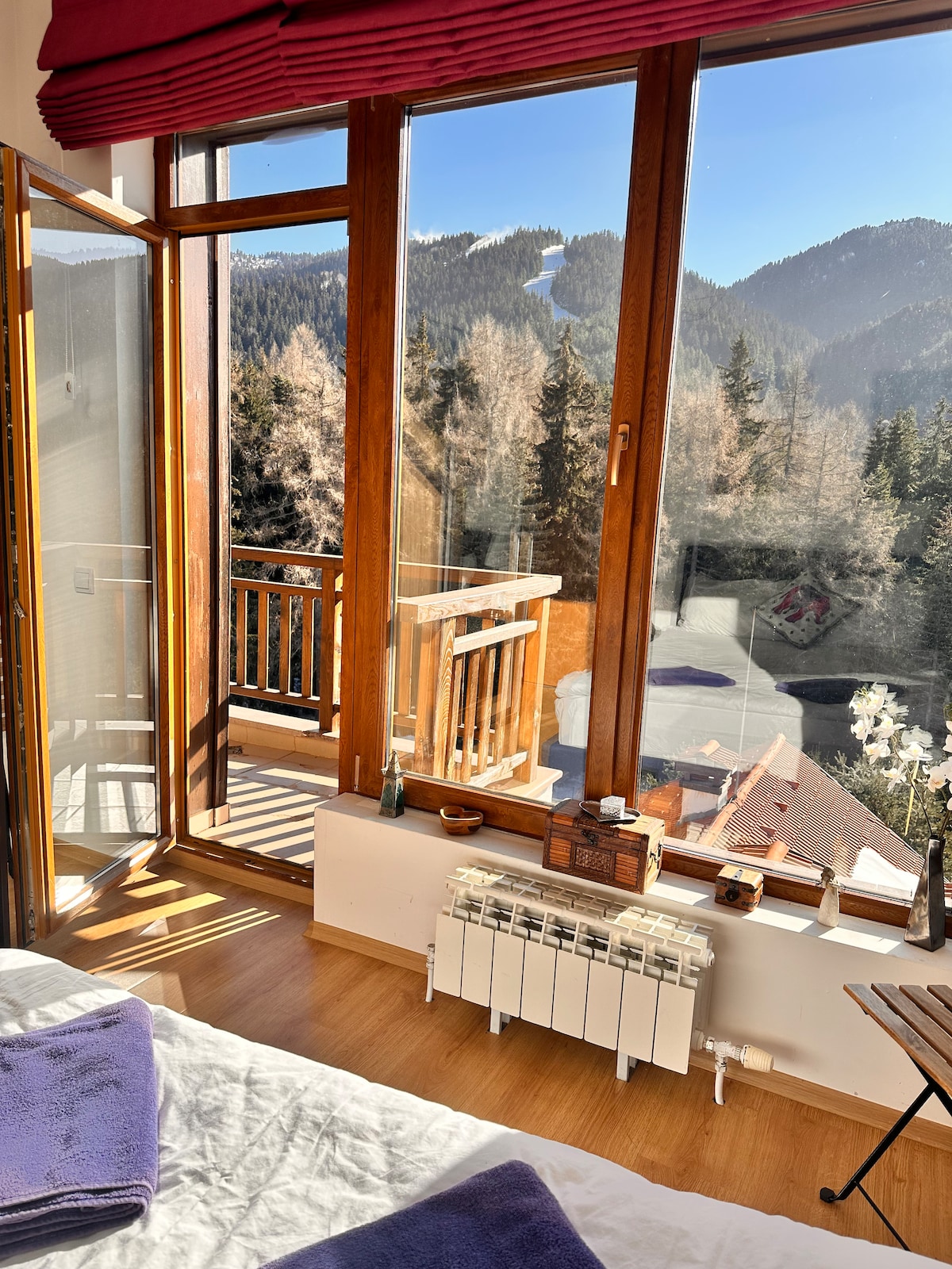 A bright bedroom features large glass doors opening to a balcony, offering views of the surrounding pine forest and mountain slopes. Natural light fills the space, highlighting the warm wooden flooring and inviting atmosphere. A cozy bed is prepared with fresh linens and purple towels.